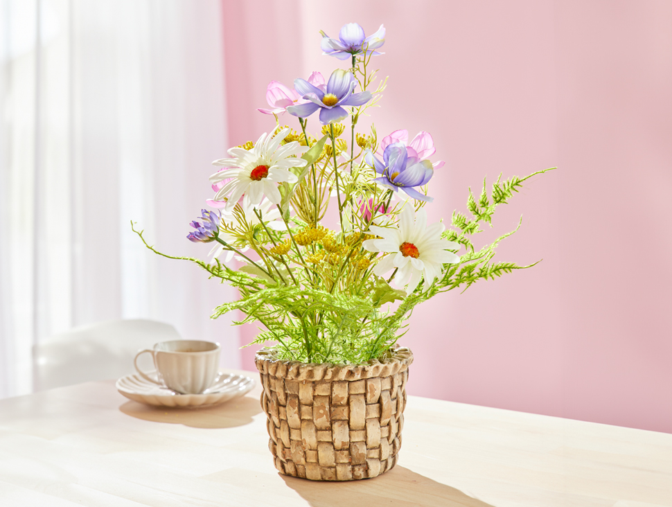 Dekorative Kunstblumen in geflochtenem Korb auf Tisch vor rosa Wand