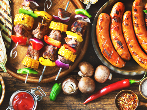 Gegrillte Spieße, Würstchen und Beilagen mit Gemüse und Gewürzen auf Holztisch