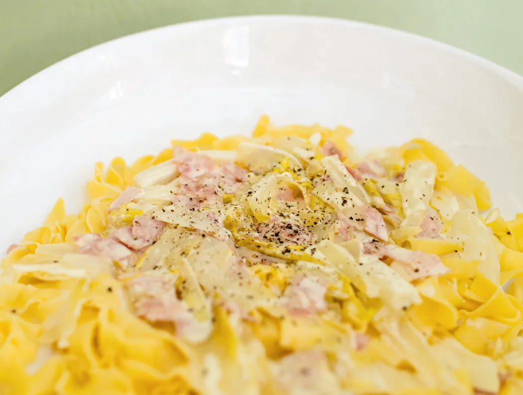 Tagliatelle mit Sahnesoße, Schinken und Käse auf einem weißen Teller.