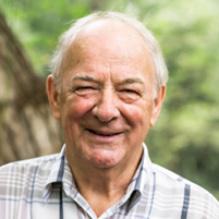 Lächelnder älterer Mann im karierten Hemd vor grünem Naturhintergrund.
