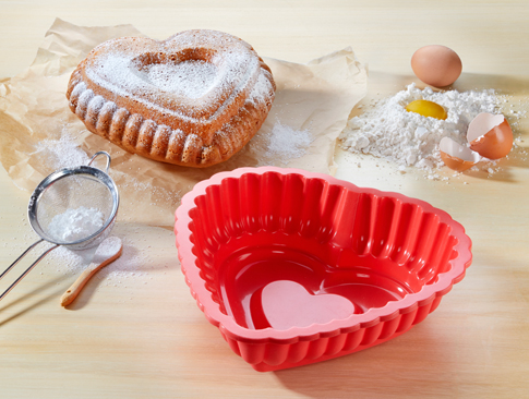 Rote Herzbackform mit gebackenem Kuchen und Backzutaten auf Holztisch.