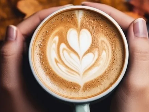 Tasse Cappuccino mit Latte Art in Herzform, gehalten von zwei Händen