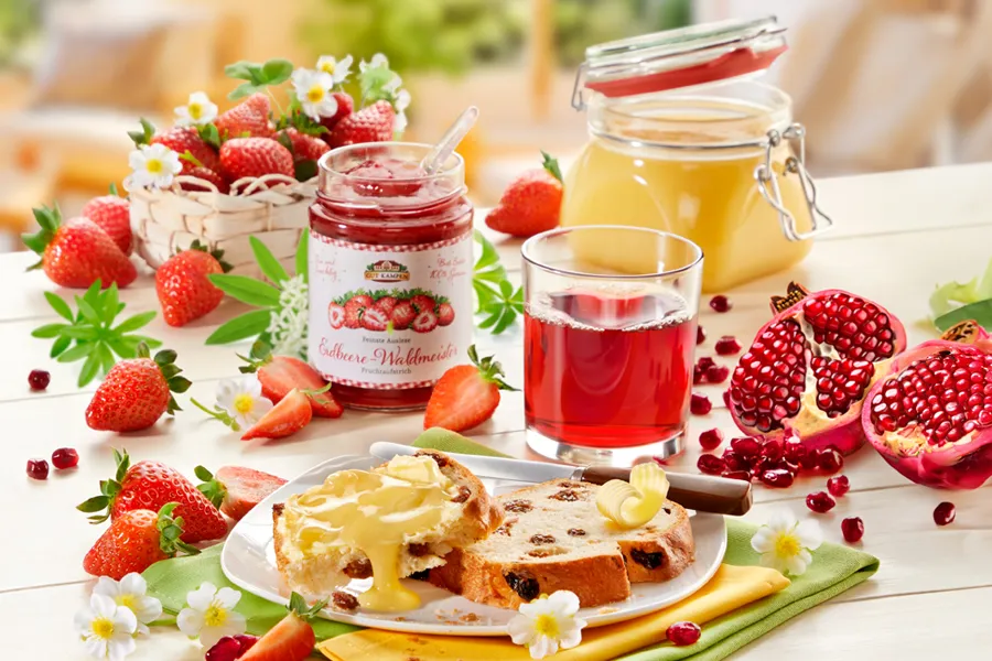 Frühstück mit Erdbeermarmelade, Saft, Granatapfel und Hefezopf auf gedecktem Tisch