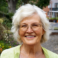 Lächelnde ältere Dame mit Brille in grüner Jacke vor blühendem Garten.